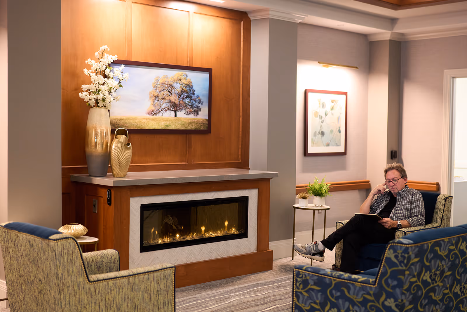 A cozy living room area with a modern fireplace set in a wooden mantel. Above the fireplace is a framed picture of a tree in a field. To the left of the fireplace is a tall vase with white flowers and a decorative gold vase. A man is sitting in a blue armchair on the right side, reading a book. The room has soft lighting, a small side table with a plant, and framed artwork on the wall.