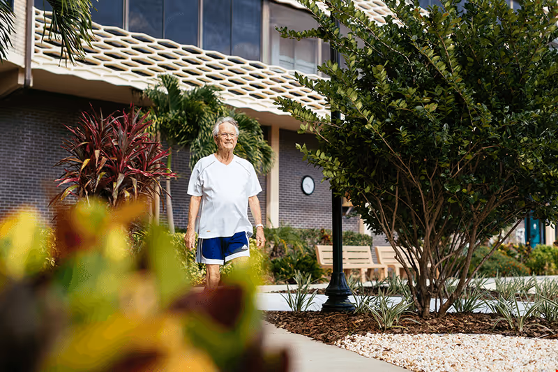 An elderly person wearing a white t-shirt and blue shorts walking on a paved path in a garden area outside a building. The surroundings include various plants, trees, benches, and a lamppost, with the building facade visible in the background.