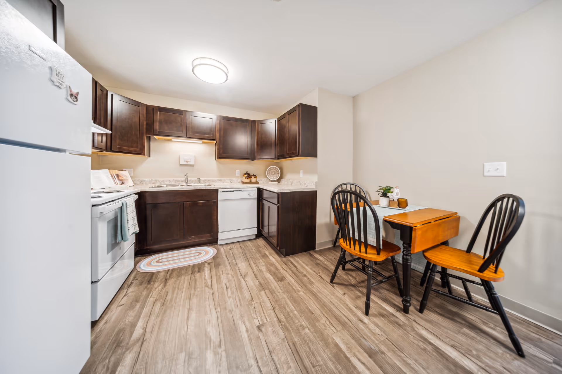 A compact kitchen area with dark wood cabinets, a white refrigerator, stove, and dishwasher. There is a small wooden dining table with two chairs on the right side, and the floor has a wood-like finish.