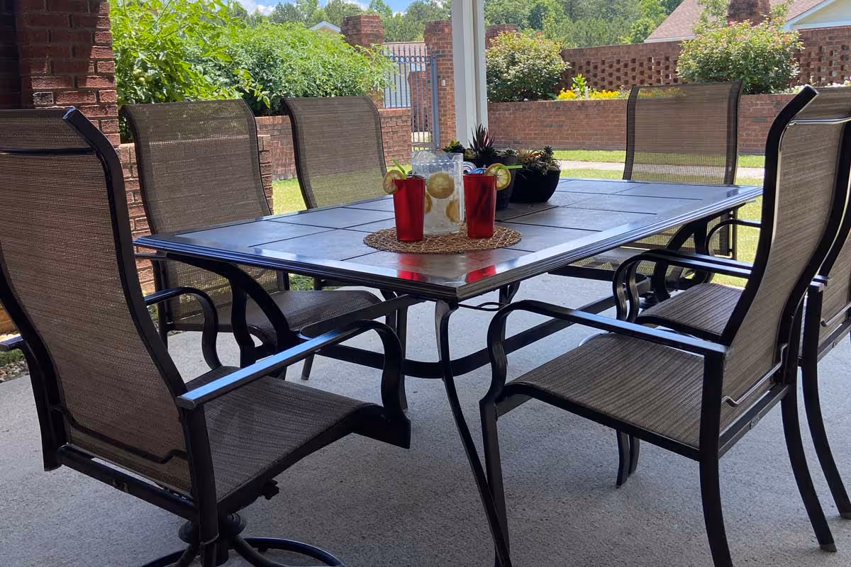Outdoor patio area with a rectangular metal table surrounded by six mesh chairs. On the table, there is a pitcher of lemon water and two red cups with lemon slices, along with a small potted plant. The patio is covered and overlooks a garden with green bushes and a brick wall.