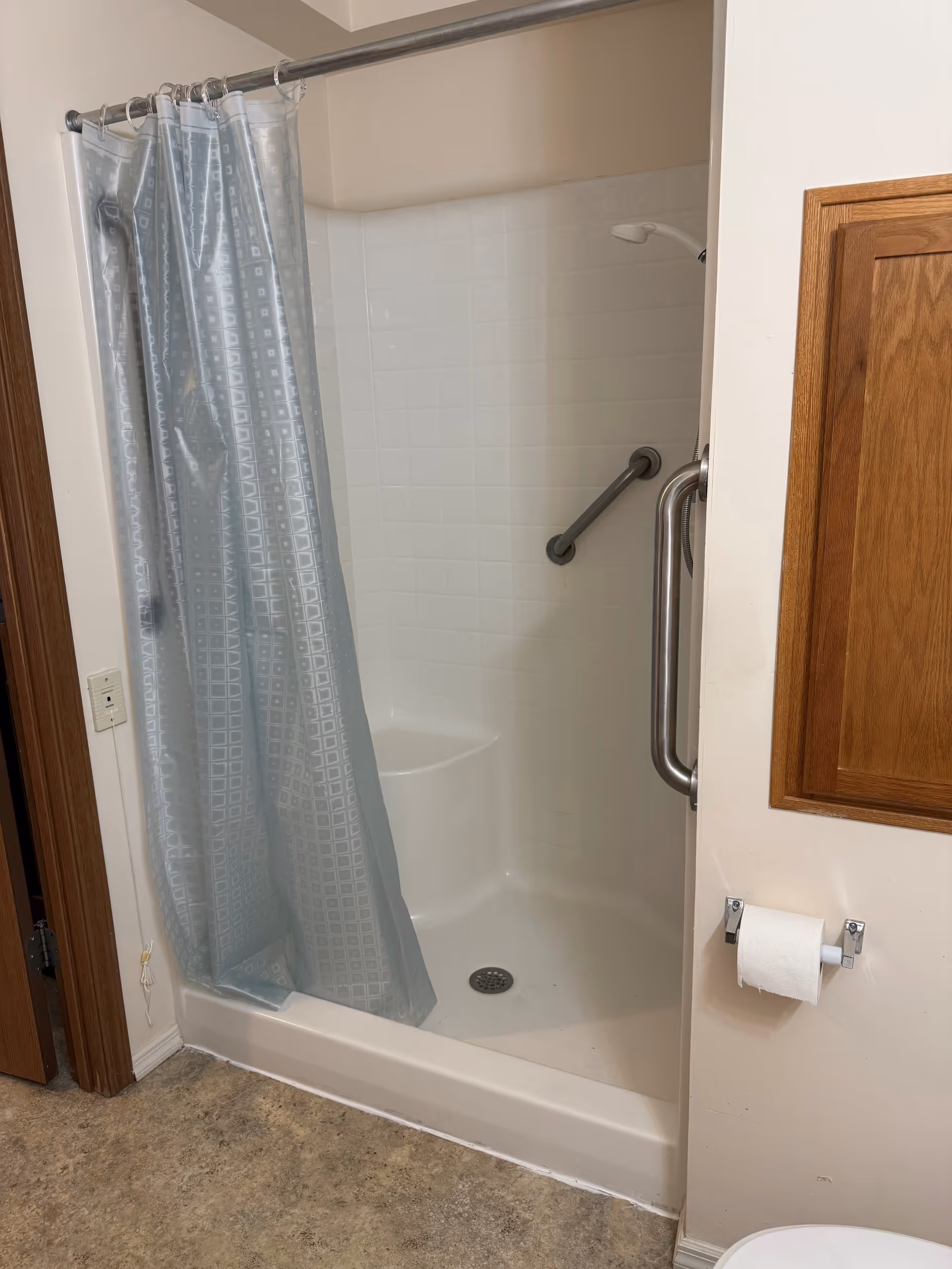 A bathroom shower area with a white tiled surround and a built-in seat. There is a silver grab bar on the wall and a silver showerhead. A light blue shower curtain with a square pattern is partially drawn. To the right, there is a wooden cabinet mounted on the wall and a toilet paper holder with a roll of toilet paper. The floor has a textured, stone-like appearance.