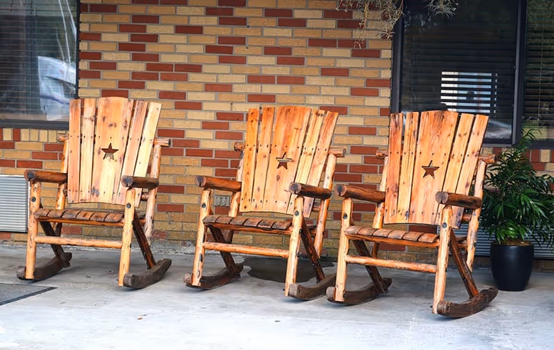 Three wooden rocking chairs with star cutouts on the backrests arranged on a concrete porch in front of a brick wall with a window and a potted plant nearby.