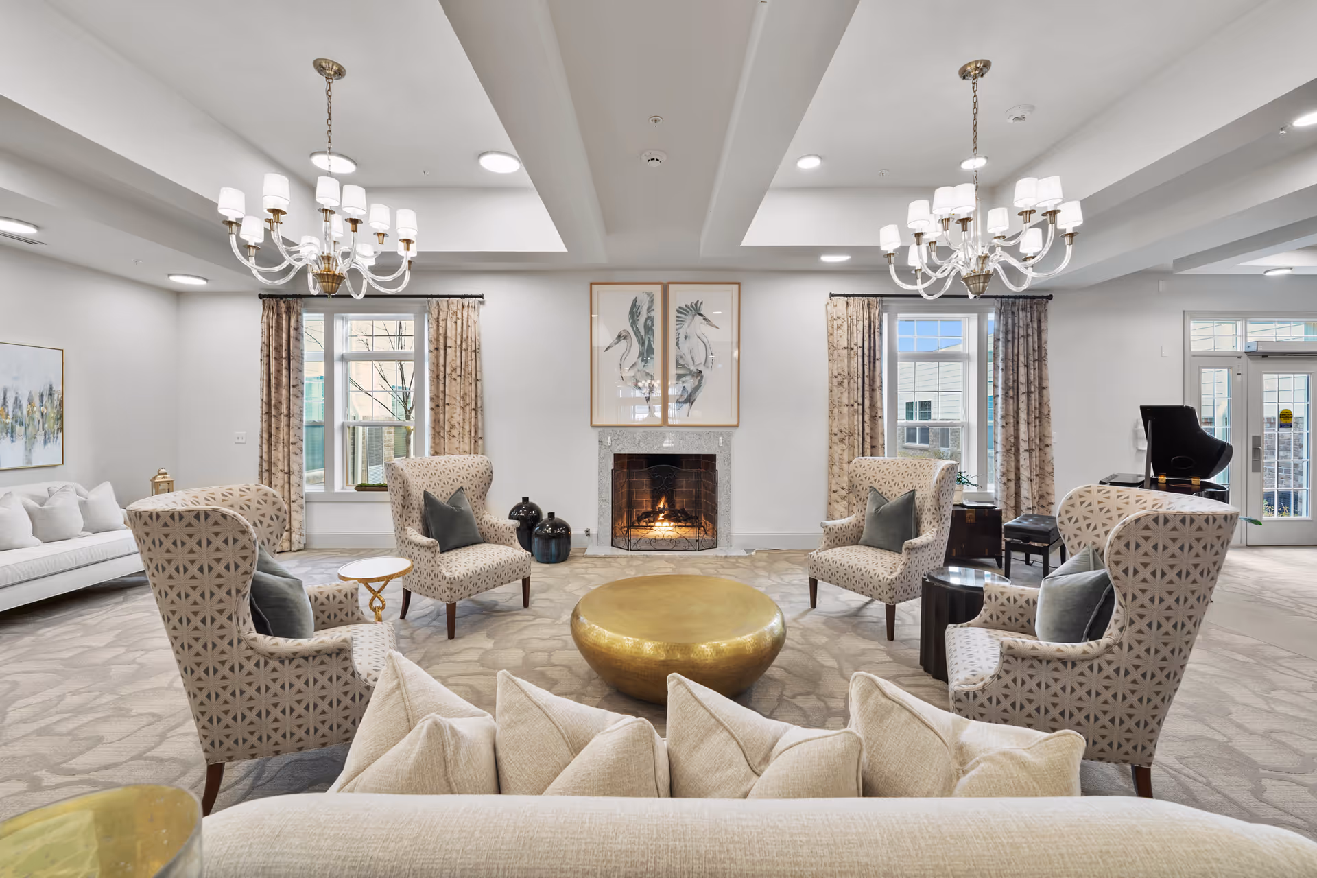 Spacious, elegantly furnished common lounge with patterned armchairs and sofas arranged around a central round gold coffee table and a lit fireplace beneath framed artwork.