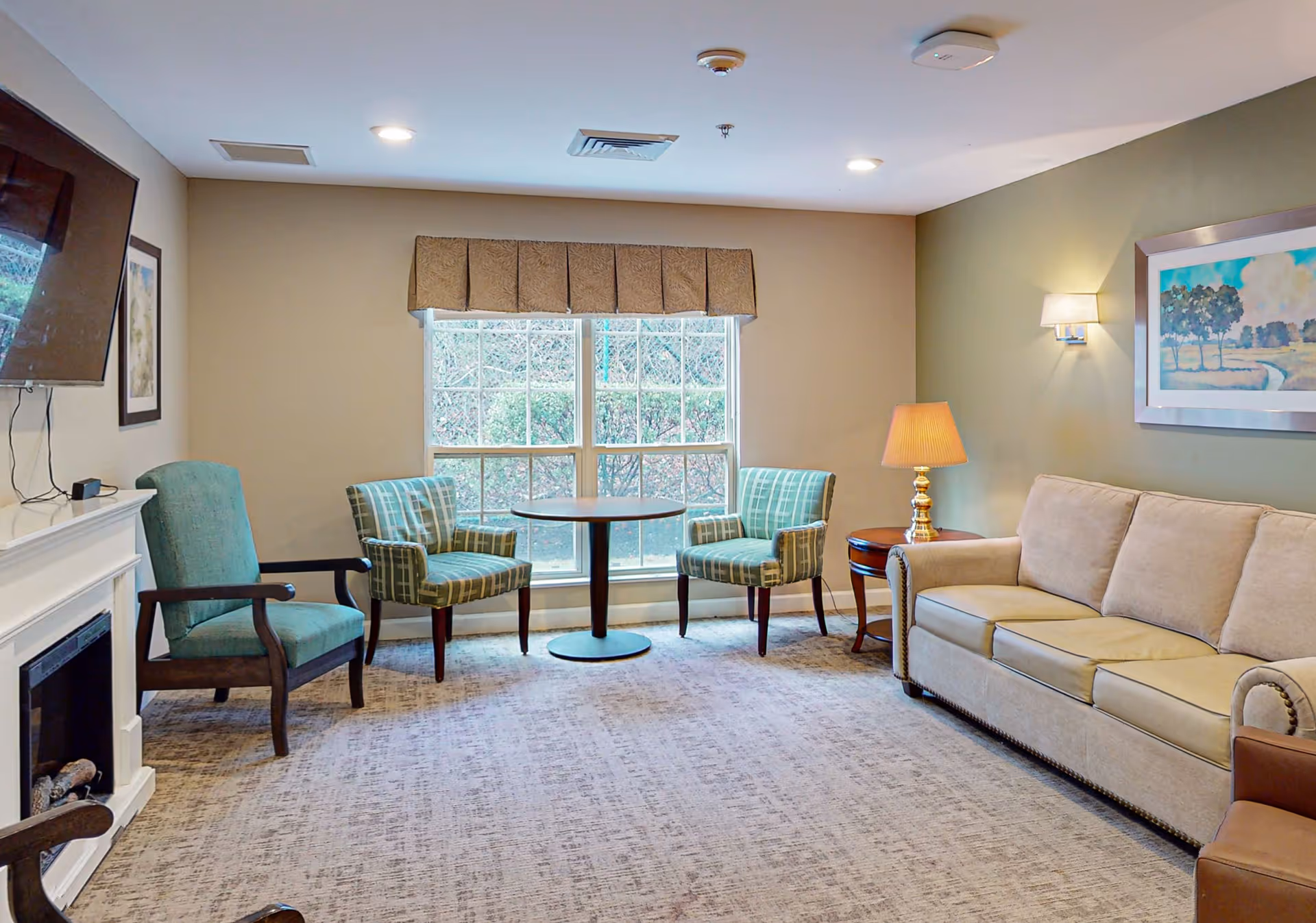 A cozy living room with a beige sofa, two green plaid armchairs, a small round table between the armchairs, a wooden side table with a lamp, a wall-mounted flat screen TV above a white fireplace, and a large window with a valance letting in natural light. The walls are painted in neutral tones and there is a framed landscape painting on one wall.