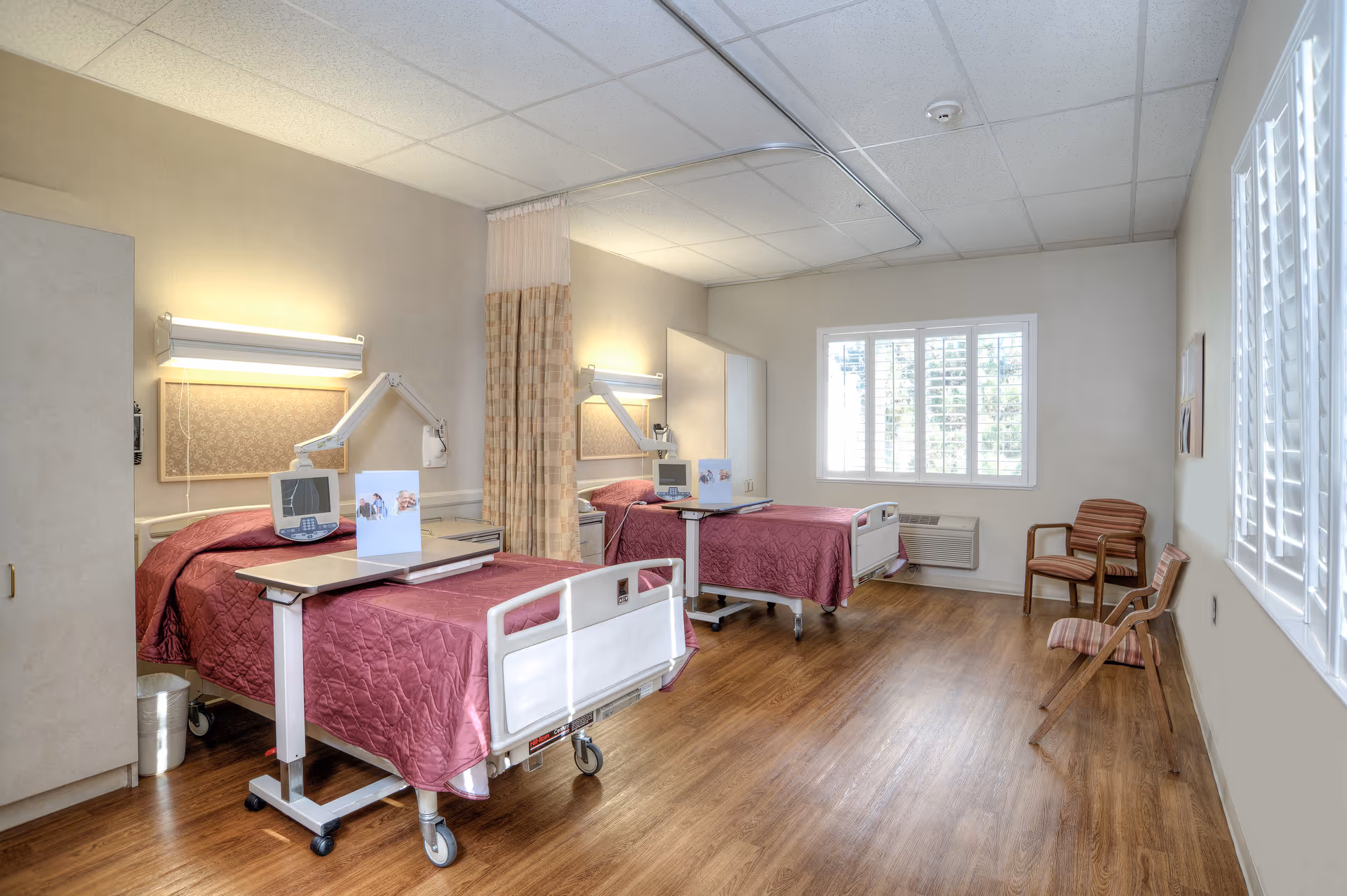 A bright and clean rehabilitation center room with two hospital beds covered in maroon quilts, each equipped with an overbed table and medical equipment. The room has wooden flooring, a large window with white shutters, two chairs against the wall, and a privacy curtain between the beds.