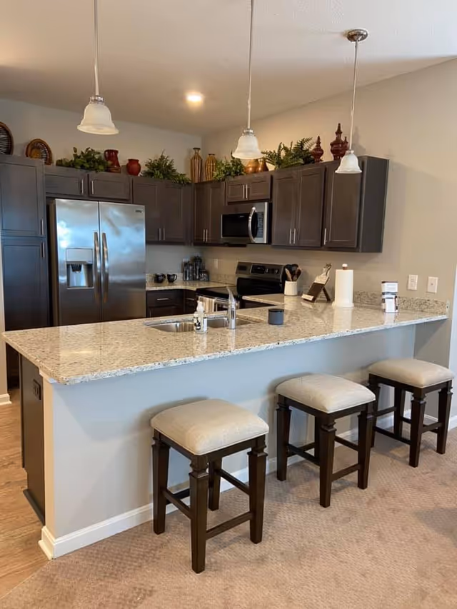 Modern kitchen with a granite countertop island featuring a sink and three cushioned stools. The kitchen has dark wood cabinets, stainless steel refrigerator, microwave, and stove. Decorative plants and vases are placed on top of the cabinets. Three pendant lights hang above the island.