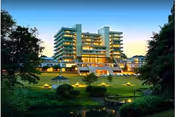 Large multi-story illuminated building at dusk with terraced lawns, a gazebo, and a reflecting pond in the foreground.
