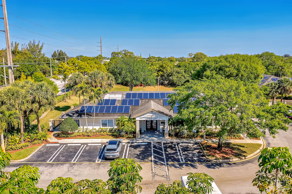 Aerial view of a single-story building with solar panels on the roof, surrounded by trees and greenery. There is a parking lot in front with one car parked, and a clear blue sky above.