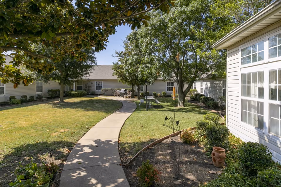 A sunny outdoor courtyard at Cedar Hill Senior Living with a curved concrete walkway, green grass, trees providing shade, and a seating area with chairs and a table near the building. The buildings have light-colored siding and multiple windows.