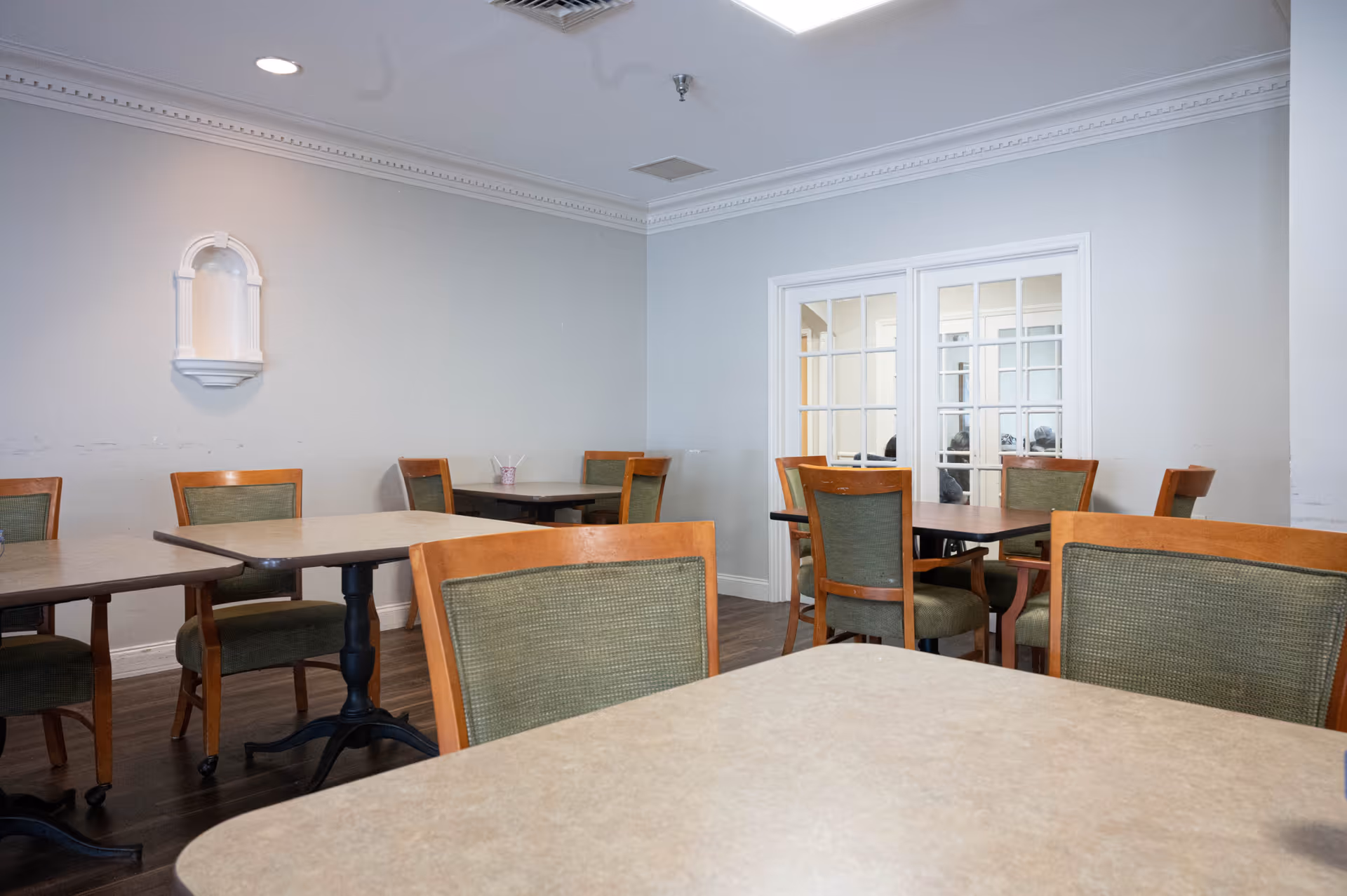 A dining area with multiple tables and wooden chairs with green cushions. The room has light-colored walls, a decorative wall niche, and a set of white-framed glass double doors. The floor is dark wood, and the ceiling has crown molding and recessed lighting.