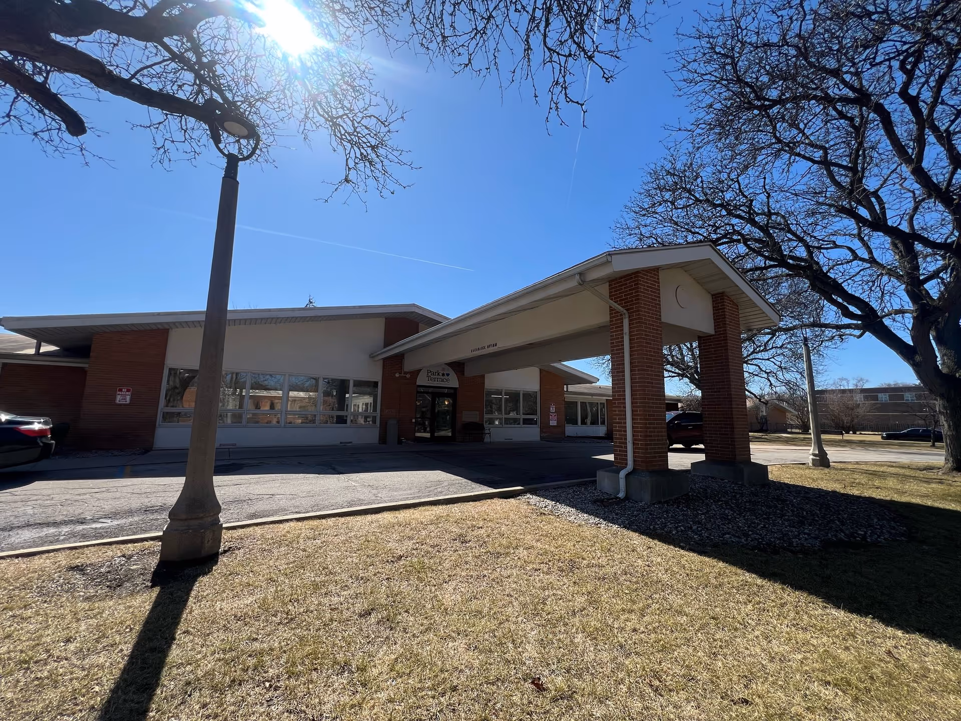 Exterior view of Park Terrace Rehabilitation Center building with a covered entrance supported by brick pillars, a lamp post, leafless trees, and a clear blue sky with the sun shining brightly.