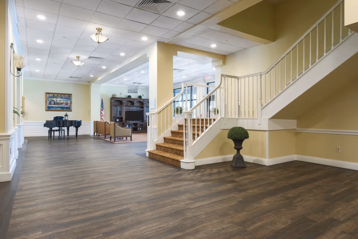 Interior view of a senior living facility featuring a staircase with carpeted steps and white railing, a wooden floor, a grand piano against the wall, a seating area with chairs and a large wooden entertainment center with a TV, and soft yellow walls with white trim.