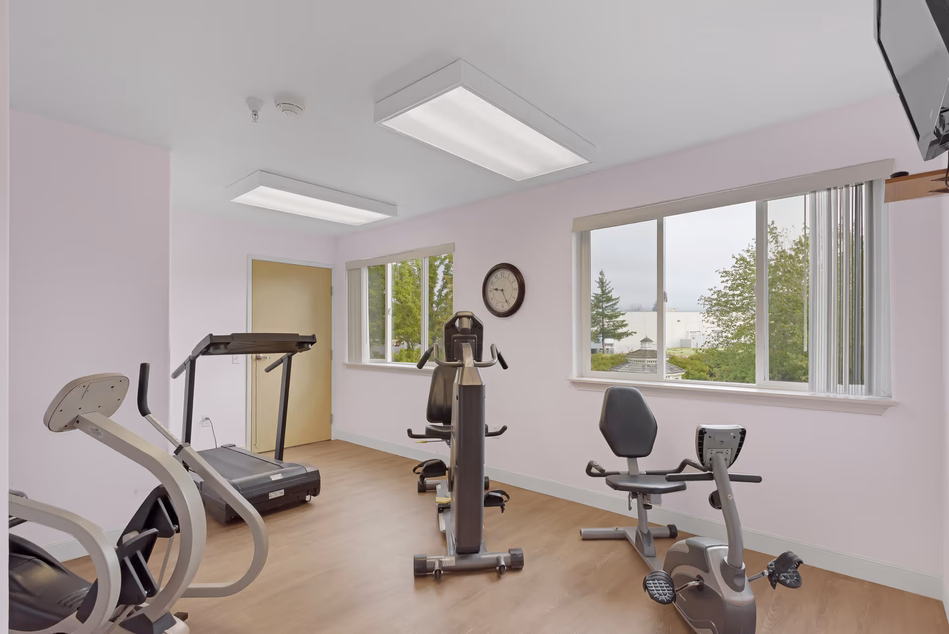 A small fitness room with exercise equipment including a treadmill, a recumbent bike, and a multi-function strength training machine. The room has light purple walls, large windows showing greenery outside, wood flooring, and ceiling lights.