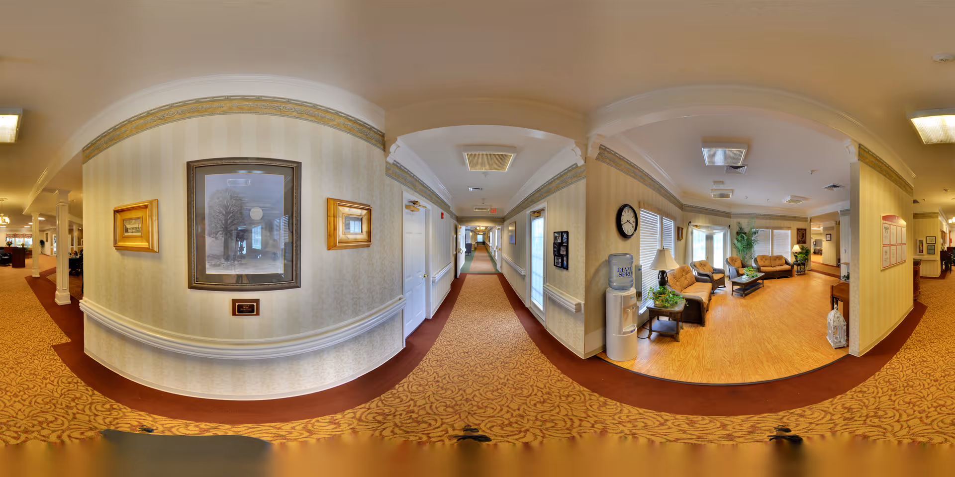 A wide-angle view of a senior living facility interior hallway with patterned carpet and wallpapered walls. The hallway leads to a sitting area furnished with sofas, chairs, a coffee table, a water cooler, and a clock on the wall. The sitting area has large windows with blinds and some plants, creating a welcoming atmosphere.