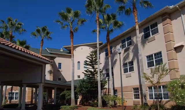 Front exterior of a three-story cream-colored senior living building with palm trees and a covered entry under a clear blue sky.