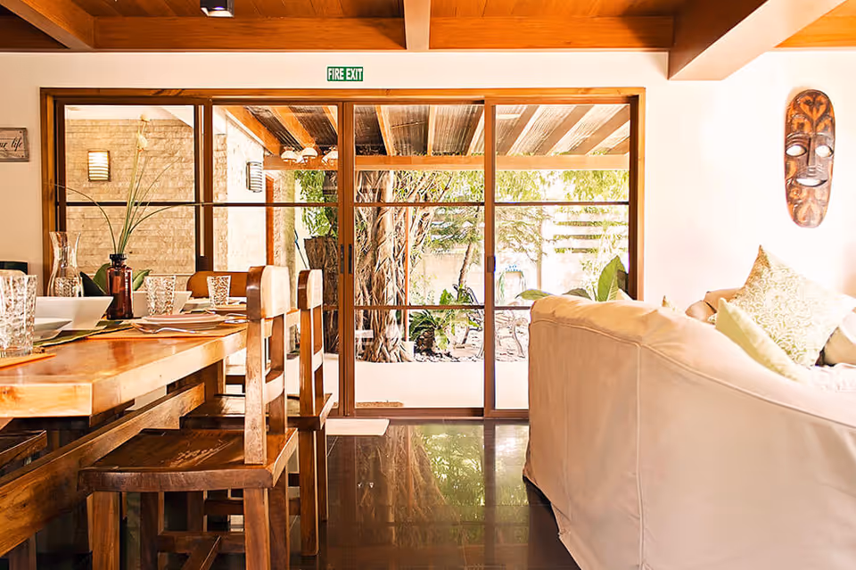 Bright open living and dining area with a wooden dining table, sofa, and large sliding glass doors opening to a garden.