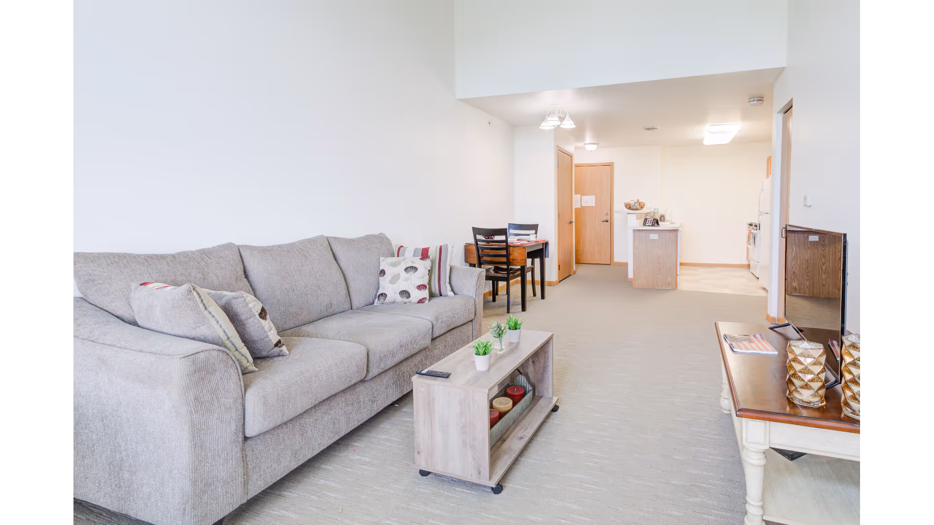 A bright living area in a senior living facility featuring a gray sofa with decorative pillows, a small wooden coffee table with potted plants and candles, a dining table with two chairs, and a kitchen area in the background with wooden cabinets and white appliances.