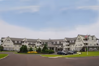 Front view of a large multi-story senior living building with a circular driveway, landscaped lawns, and a flagpole.
