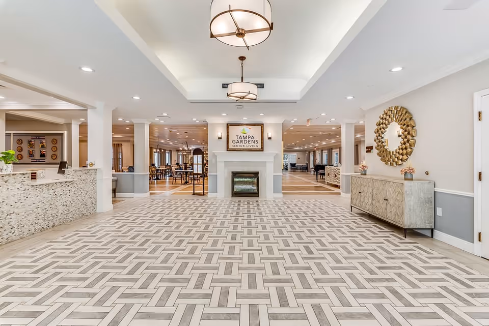 Spacious and well-lit interior of Tampa Gardens Senior Living featuring a patterned tile floor, a reception desk on the left, a decorative fireplace with a Tampa Gardens Senior Living sign above it in the center, and a sideboard with a round golden mirror on the right. The area opens into a dining space with tables and chairs in the background.