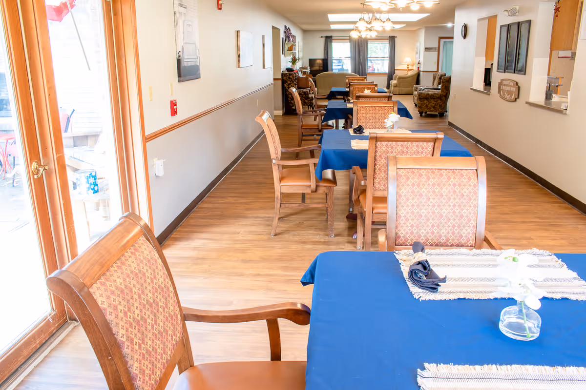 Interior view of a senior living facility dining area with wooden chairs and tables covered with blue tablecloths. The room has wooden flooring, large windows with curtains, and a seating area with armchairs in the background. There is a glass door on the left side leading outside.