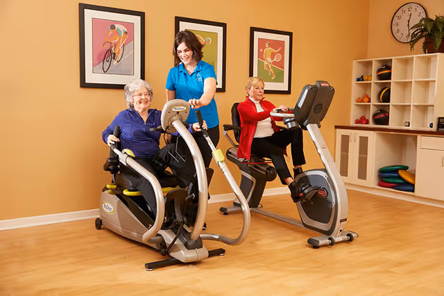 Two elderly women exercising on stationary recumbent bikes in a fitness room with a female staff member assisting one of them. The room has wooden flooring, framed sports-themed artwork on the wall, a clock, and shelves with exercise equipment.