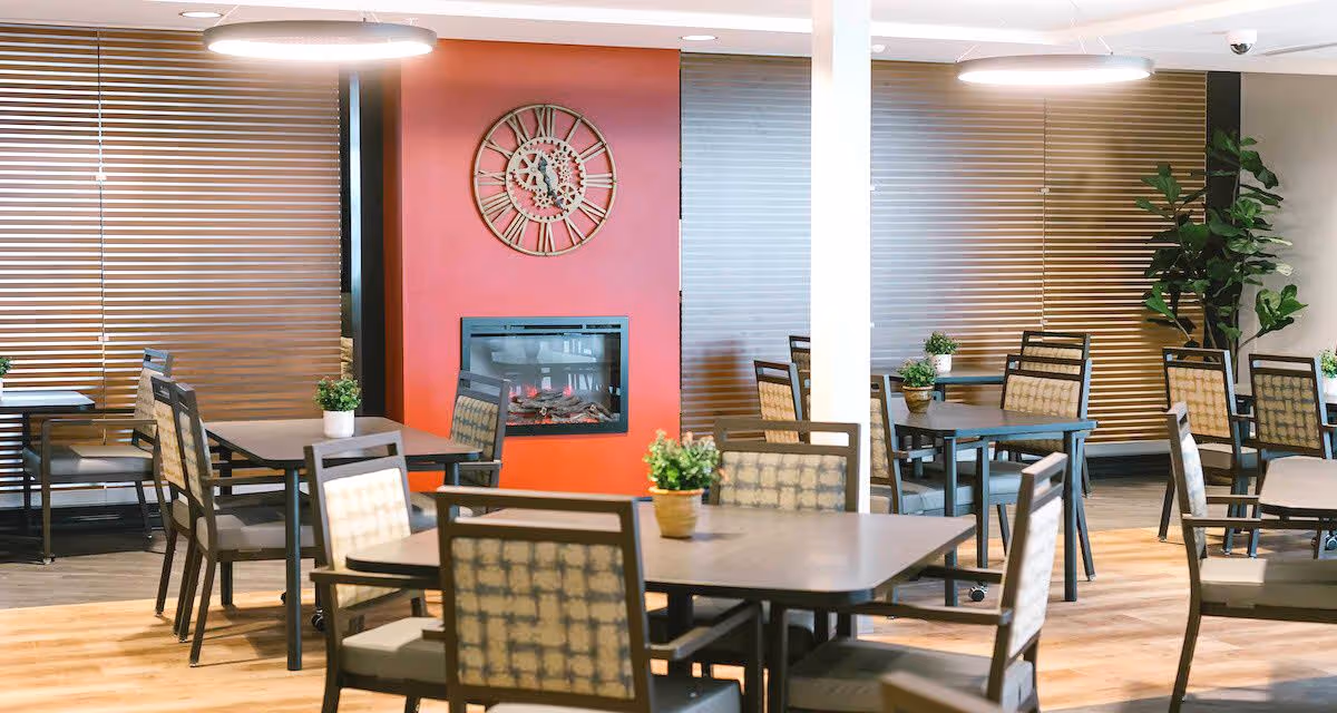 A bright dining room with multiple square tables and cushioned chairs arranged neatly. Each table has a small potted plant as decoration. The room features wooden blinds on the windows, a red wall with a large decorative clock above a modern fireplace, and a tall green plant in the corner. The floor is wooden, and the ceiling has round light fixtures.