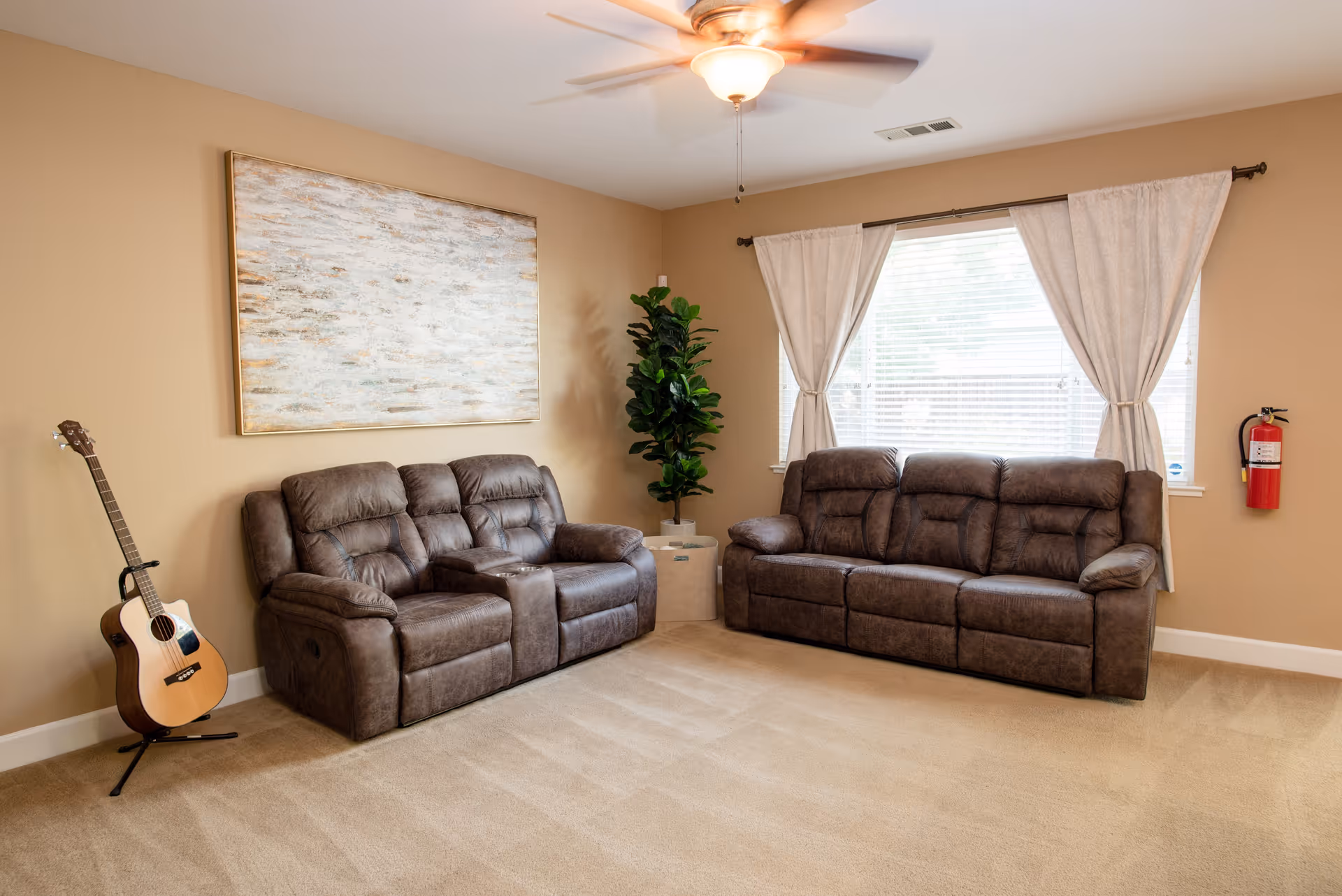 A cozy living room with two brown leather sofas arranged in an L-shape, a large abstract painting on the beige wall, a tall green potted plant in the corner, a wooden acoustic guitar on a stand, beige carpet flooring, a ceiling fan with a light, and a window with white curtains and blinds. A red fire extinguisher is mounted on the wall near the window.