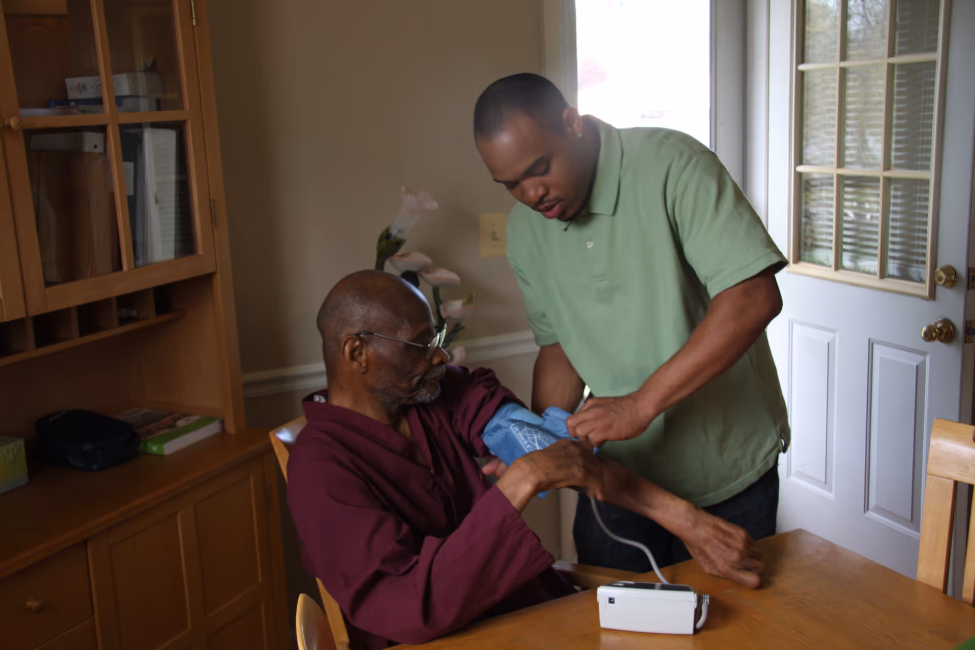 A caregiver in a green polo shirt is helping an elderly man in a maroon robe by measuring his blood pressure using a blood pressure cuff and monitor at a wooden table in a well-lit room with a glass-paneled door and wooden cabinet.