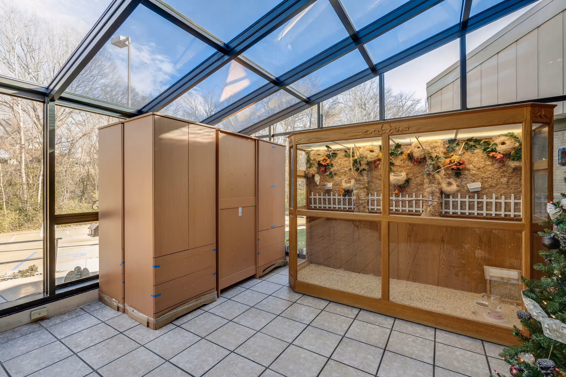 Sunlit glass-roofed atrium with tile floor containing wooden storage cabinets and a large wooden display enclosure decorated with nests and plants.