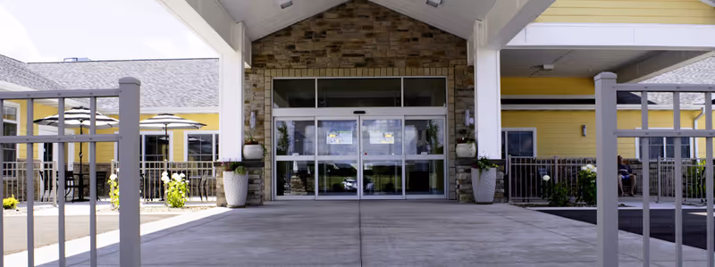 Covered entrance of a yellow assisted living building with a stone facade, sliding glass doors, planters and a paved drop-off area.