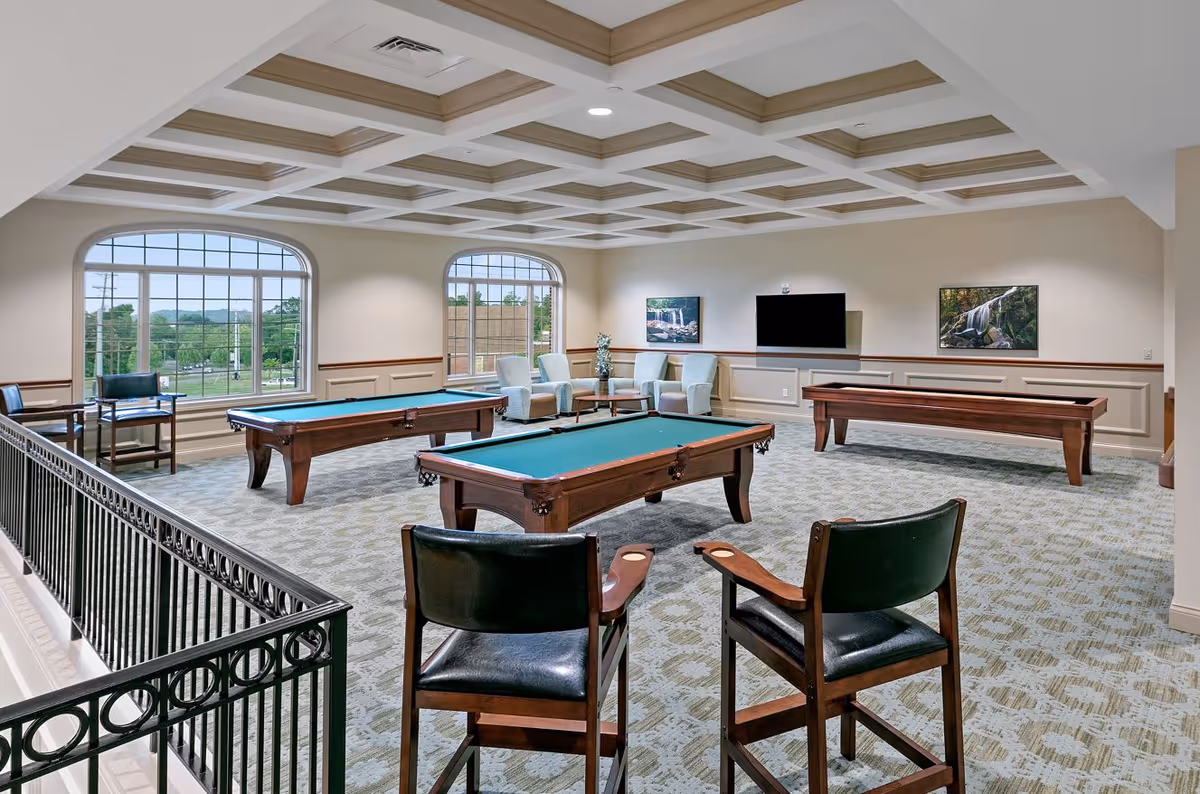 Well-lit recreation room with pool tables, shuffleboard, seating, and large arched windows.