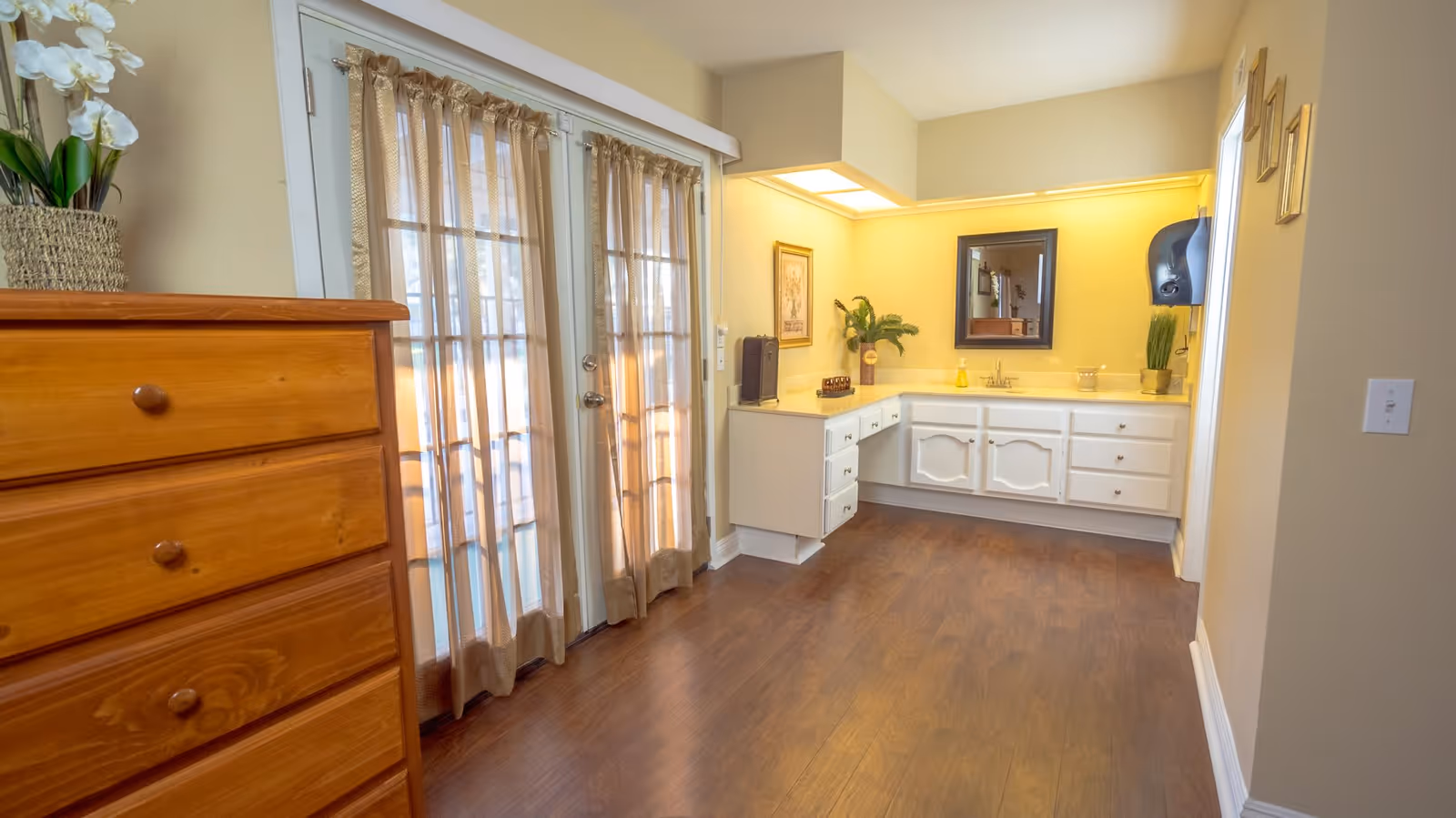 Interior view of a room with wooden flooring featuring a wooden chest of drawers on the left and a white vanity with multiple drawers and a mirror on the right. The room has beige walls, a set of glass double doors with sheer beige curtains, and decorative plants and framed pictures on the walls.