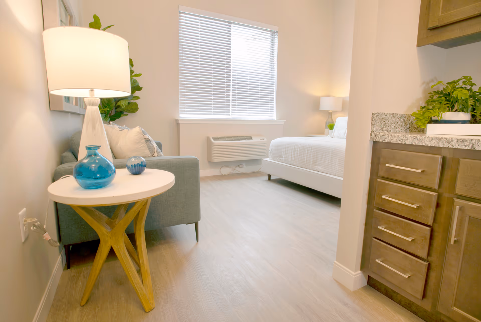 Bright assisted living studio room with a sofa and side table holding blue vases and a lamp, a bed by a window with blinds, and kitchenette cabinets.