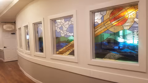 A curved hallway with wooden flooring and white walls featuring four stained glass windows depicting a colorful landscape with a sun, hills, and sky. At the end of the hallway, a door with a sign above it reads 'CHAPEL & QUIET ROOM'.