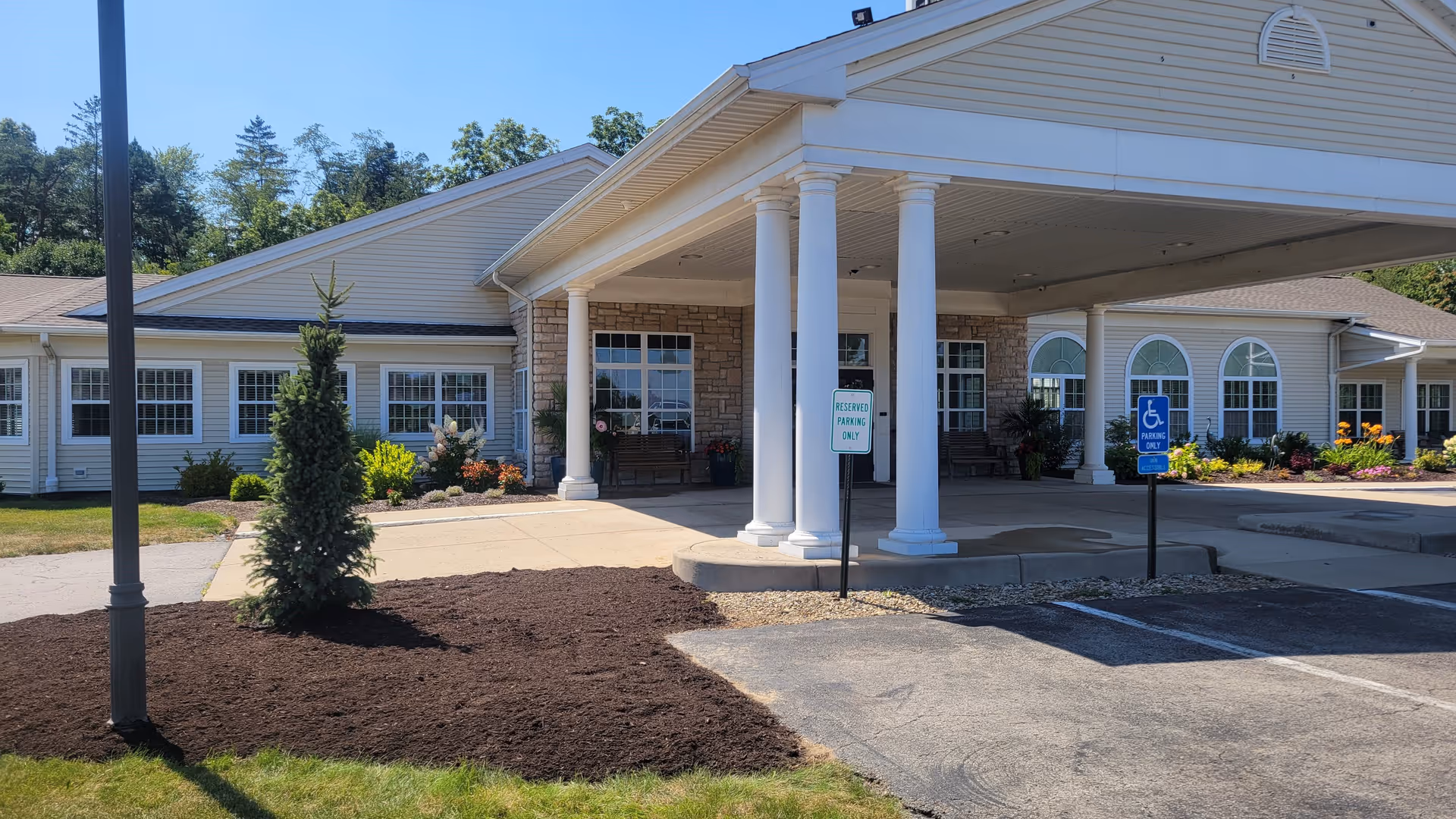 Exterior front view of Whispering Pines Village showing a covered entrance with white columns, a reserved parking sign, a handicapped parking sign, and landscaped areas with small trees and flowers under a clear blue sky.
