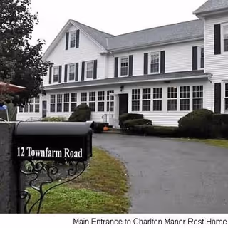 White multi-story building with black shutters and a long row of windows on the ground floor, surrounded by bushes and a driveway. A black mailbox in the foreground displays the address '12 Townfarm Road'.