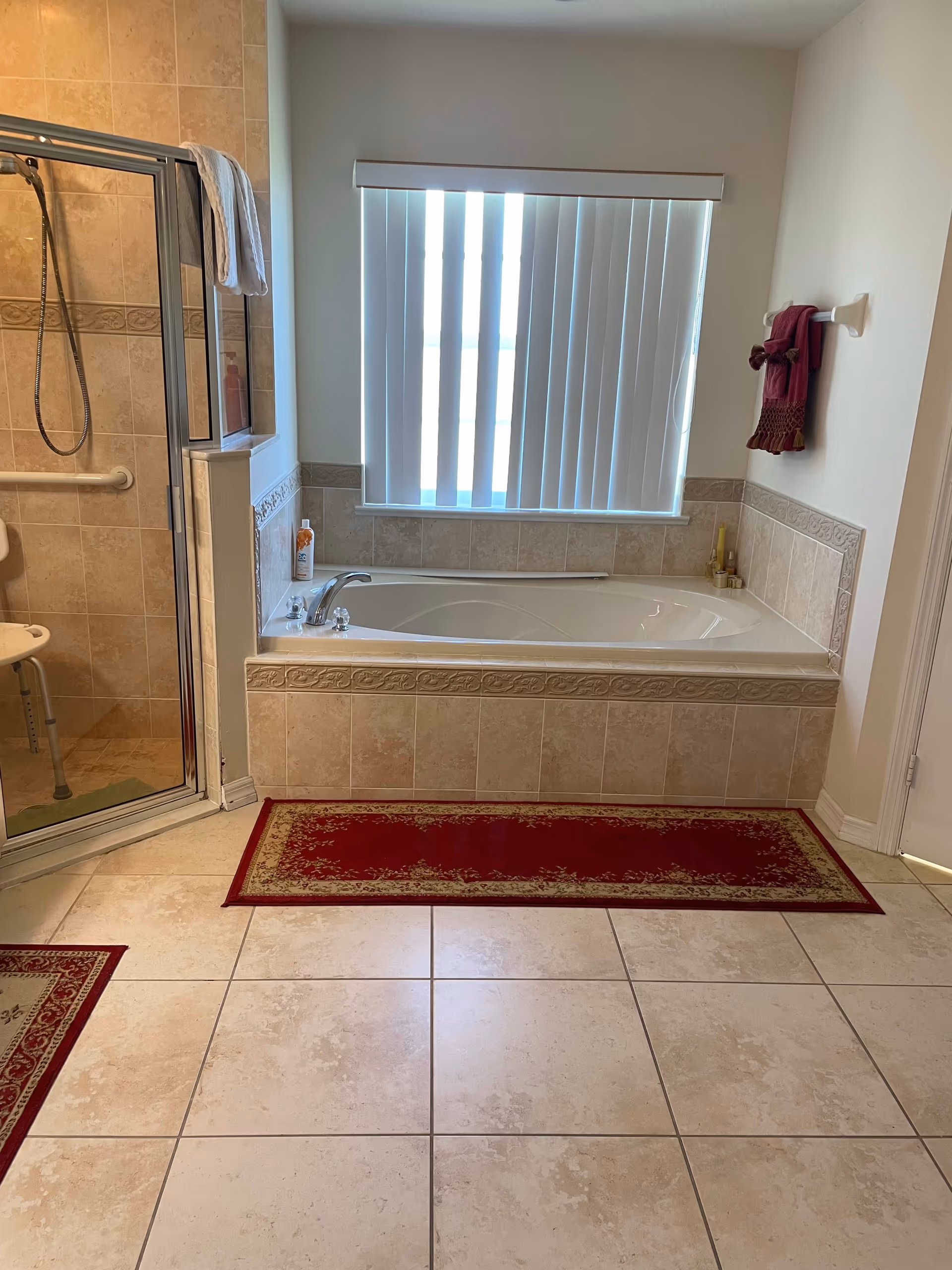 A bathroom with a large bathtub surrounded by beige tiles and a window with vertical blinds above it. To the left, there is a glass-enclosed shower with a white towel hanging on the door and a shower chair inside. The floor is tiled with light-colored tiles, and there are two red and gold patterned rugs on the floor. A towel rack with a red towel is mounted on the wall to the right.