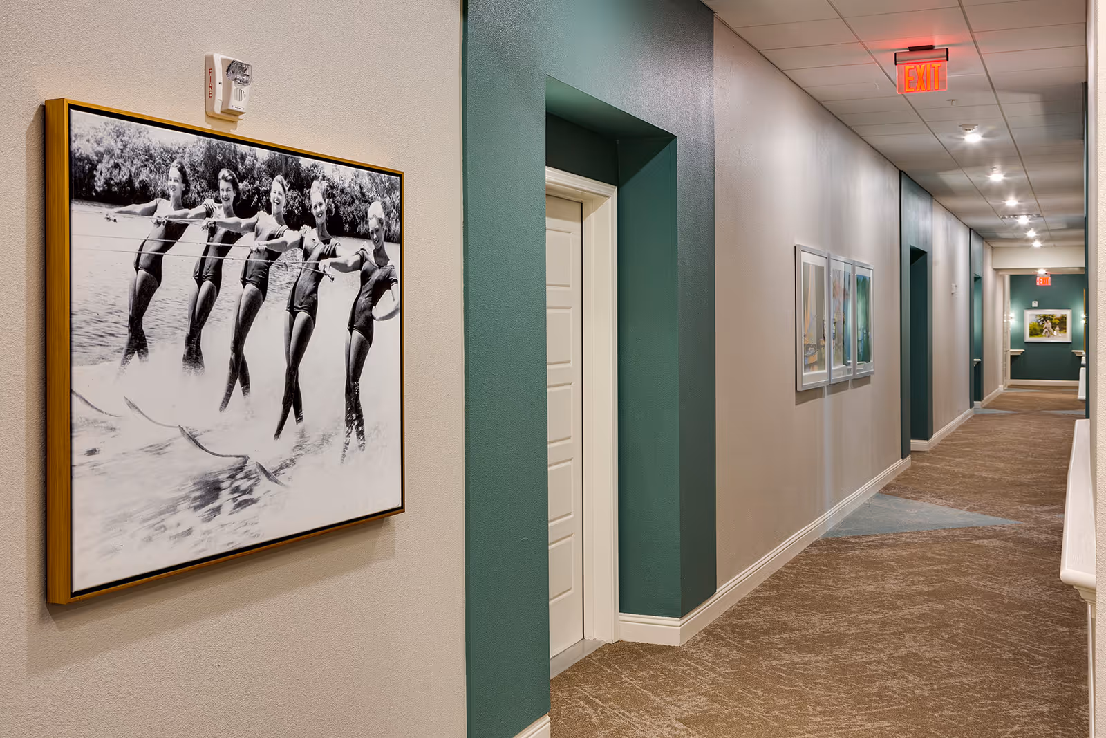 A well-lit interior hallway with beige walls and carpeted floor. The hallway features several doorways framed with green accents and is decorated with framed pictures on the walls, including a large black and white photo of six women water skiing. Ceiling lights and an exit sign are visible at the far end of the corridor.
