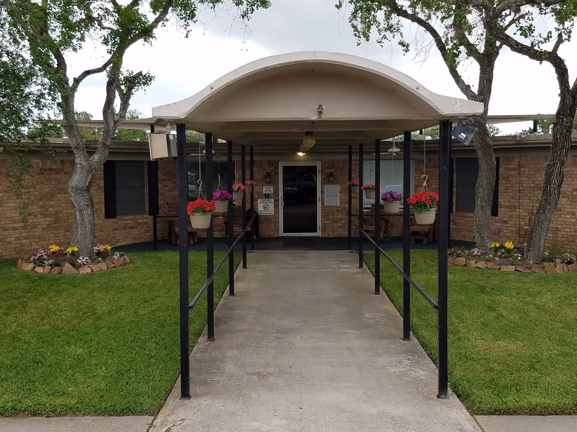 Entrance to a brick building with a covered walkway supported by black metal poles. There are hanging flower pots with red and pink flowers along the walkway. The building has windows with black shutters and flower beds with colorful flowers on either side of the entrance. Trees are visible around the building.