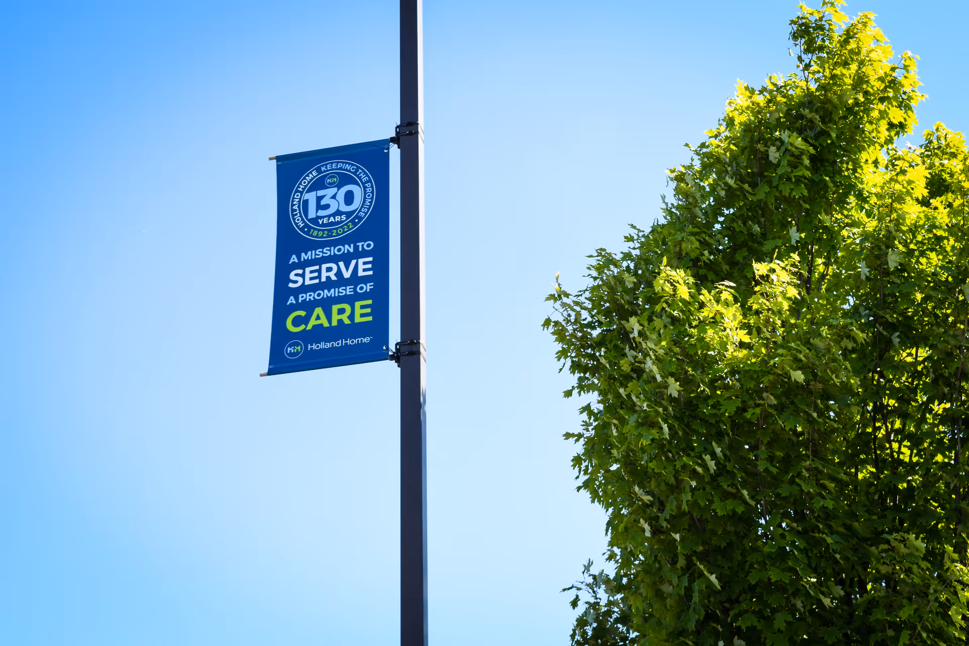 A blue Holland Home street banner on a lamppost beside a leafy green tree against a clear blue sky.