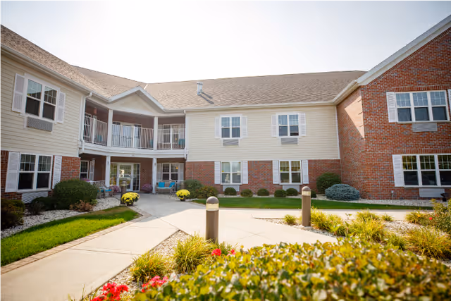 Courtyard and front entrance of a two-story assisted living building with walkways, landscaping, and outdoor seating.