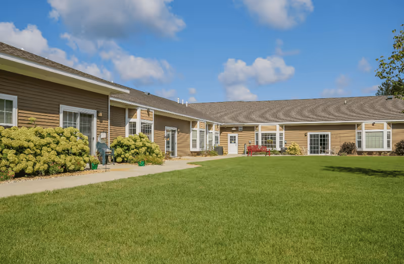 Exterior view of a single-story assisted living facility building with brown siding, multiple windows, and a well-maintained green lawn in front under a partly cloudy blue sky. There are bushes and a red bench near the building.