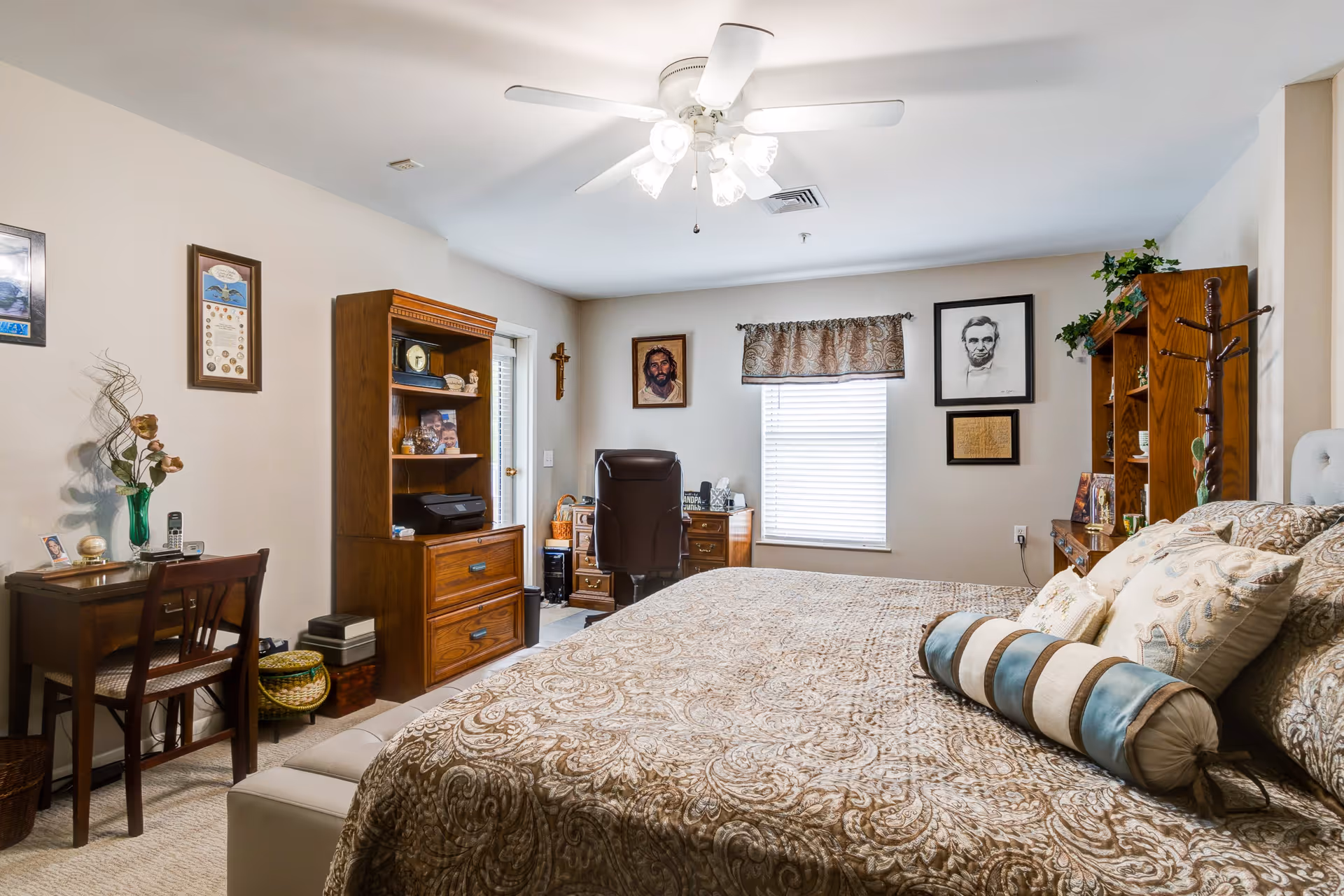 A cozy bedroom with a large bed covered in a patterned beige and brown bedspread with multiple pillows. The room features wooden furniture including a desk with a chair, a bookshelf, and a dresser with a black office chair. There are framed pictures and artwork on the walls, including portraits of Jesus and Abraham Lincoln. A ceiling fan with lights is mounted on the ceiling, and a window with a decorative valance lets in natural light.