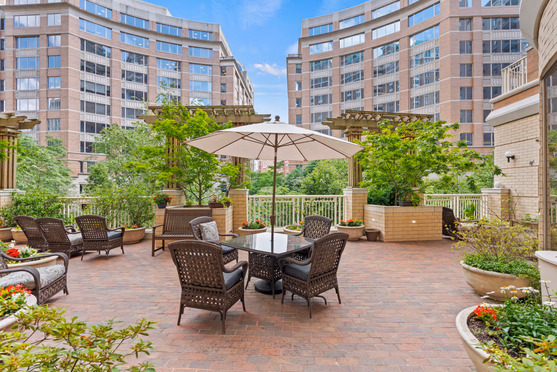 Outdoor patio area with wicker chairs and a table under a large umbrella, surrounded by potted plants and greenery. In the background, there are tall residential buildings under a partly cloudy sky.
