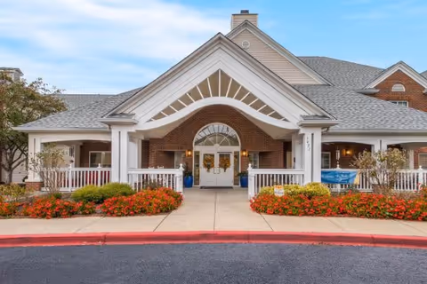 Entrance of a brick senior living building with a white covered portico, double doors, and flowerbeds in front.