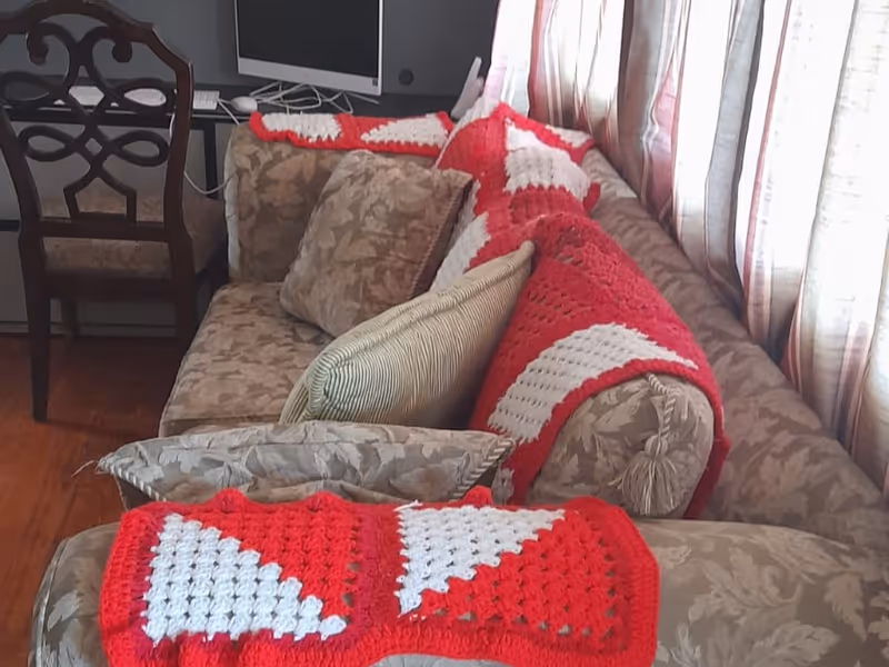 Floral upholstered sofa in a living room with red-and-white crocheted blankets draped over it, cushions, a chair and a TV in the background.