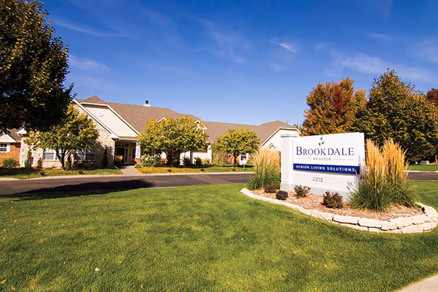 Exterior view of Brookdale Brighton senior living facility on a sunny day with a clear blue sky, green lawn, trees with autumn foliage, and a sign in the foreground that reads 'Brookdale Brighton Senior Living Solutions 2215'.