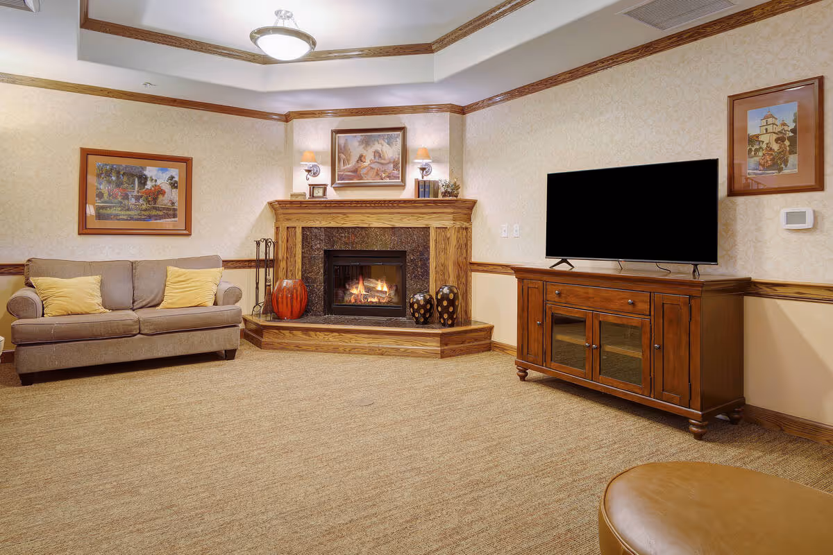 A cozy living room with a beige sofa adorned with two yellow pillows, a wooden fireplace with a fire burning, decorative vases on the hearth, a flat-screen TV on a wooden cabinet, and framed artwork on the walls.