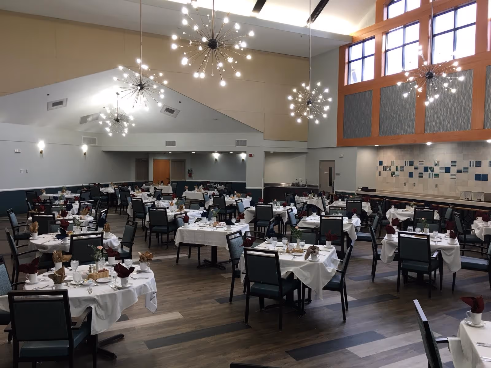 A spacious dining room with multiple tables covered in white tablecloths, each set with cups, glasses, napkins, and small centerpieces. The room features modern, starburst-style chandeliers hanging from a high ceiling, large windows letting in natural light, and a decorative tiled wall on one side.