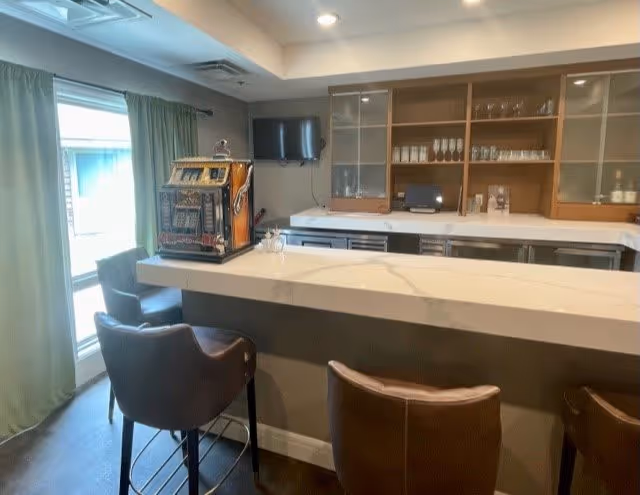 Interior view of a senior living facility's bar area with a marble countertop, three brown leather bar stools, a vintage slot machine on the counter, a wall-mounted TV, and wooden cabinets with glassware behind the counter. A window with green curtains is visible on the left side.